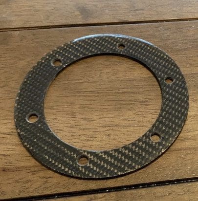 Carbon fiber circular gasket with six holes on a wooden surface.