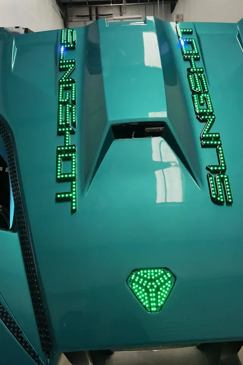 Close-up of a teal sports car hood with green LED lights and a glowing emblem.